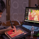 A young woman wearing headphones uses a stylus on an art program on a tablet while a laptop in front of her shows the same image.